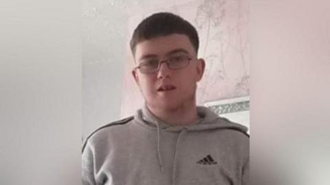 Daniel Ambler, a 23-year-old man with short dark hair, wearing spectacles and a grey hoodie. He is standing in what looks like a living room. There is pink and grey wallpaper on the wall behind him.
