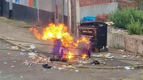 A wide shot of Sylvester Road in the Vauxhall area of Liverpool. There is a purple bin on fire surrounded by litter strewn across the pavement and road.