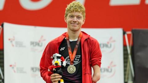 Para-badminton athlete Rob Donald stands proud with a silver medal around his neck. He is smiling and holding what appears to be a dog mascot, that is saluting, in one hand. 