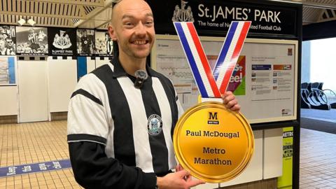 Paul McDougal stands in front of St James' Park Metro station wearing Newcastle football black-and-white striped kit. He is holding up a large paper medallion which says he has completed a Metro marathon and credits his name. Paul has a shaved head and stubble. 