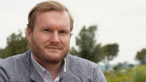 Alexander Sparrow sits in front of trees on a cloudy day. He is wearing a grey jumper with a white collared shirt underneath. He has short hair with a dark, neatly-trimmed beard.