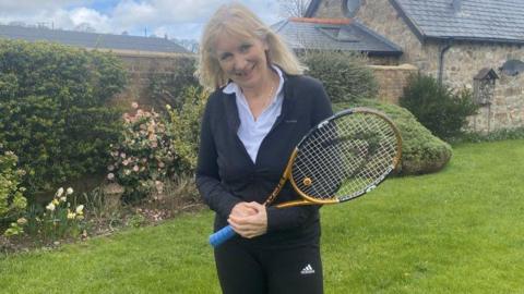 A woman with blonde straight hair, a black sports outfit is holding a yellow and blue tennis racket and standing in a garden