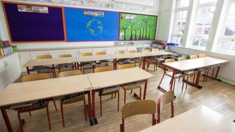 An empty classroom with tables and chairs in the foreground and a wall display in the background