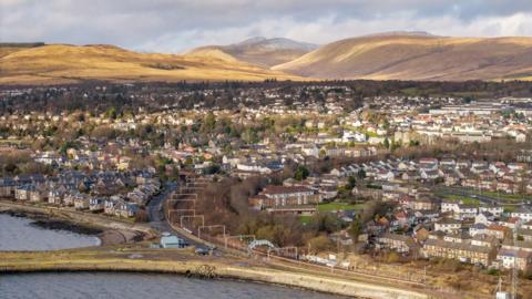 A town on the coast with lots of houses and trees and a road running along the water. Mountains can be seen just beyond the boundary of the town
