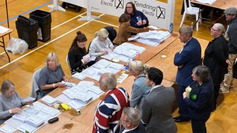 Vote counting in Tendring in Essex. 6 tellers sit at a table counting votes while candiates in suits, one of them wearing a union flag suit watch on