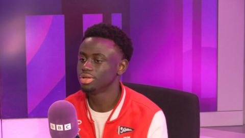 Dolapo Okunola has short dark hair, we wears a red and white jacket and speaks into a purple BBC microphone