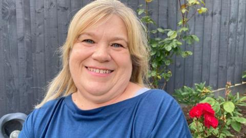 Janet Wilcock, who has blonde hair and is wearing a blue top, smiles at the camera in her garden