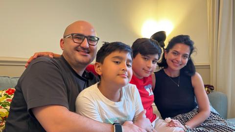 Akmal is wearing a dark grey top and wearing black square glasses, Gafur is wearing a white t-shirt, Bobur is wearing a red t-shirt and Gavhar is wearing a black vested top and a black and white checkered skirt and sitting on a blue sofa. 