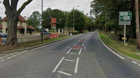 A two‑lane road stretching into the distance, bordered by trees and greenery on both sides. On the right side of the road, there are directional road signs mounted on a pole, indicating routes toward nearby places. A wooden fence and a grassy verge run alongside the road in that direction. On the left side, there is a building with a red roof set behind a wooden picket fence, with several cars parked nearby. The road markings include central white dashed lines and red rectangular patches designed to slow traffic.