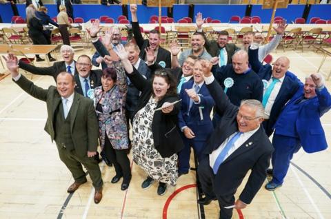 Reform candidates celebrate the Hartlepool election result. There are a group of 16 men and woman standing close together in a group with their hands raised in the air and celebrating. Many of them are wearing light blue and white rosettes. They are standing in a leisure centre with markings on the floor and a row of desks and chairs behind them