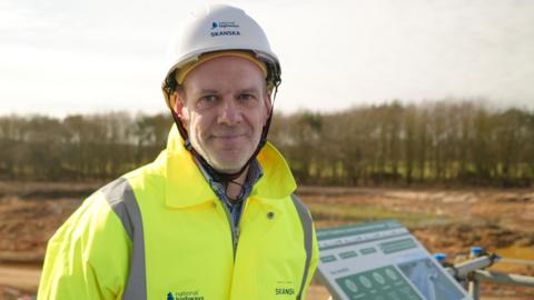 Alistair Stout is wearing a hard hat branded with the word Skanska and is also wearing a high-vis yellow jacket. He is smiling and standing in front of the construction site.