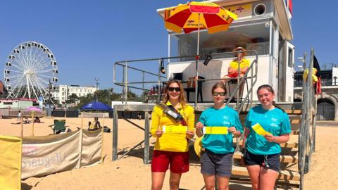 Bournemouth Beach with RNLI lifeguard tower behind a lifeguard and council beach safety staff holding yellow wristbands.