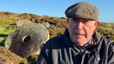 Jude McHugh - A man sitting outside with a mill stone in the background behind him. He is wearing a navy jacket and a cap.