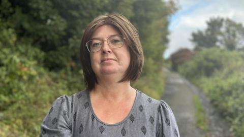 Elaine O'Boyle is standing on a country path, with trees and bushes on either side. She is wearing a grey patterned top, has a brown bob and is wearing glasses.