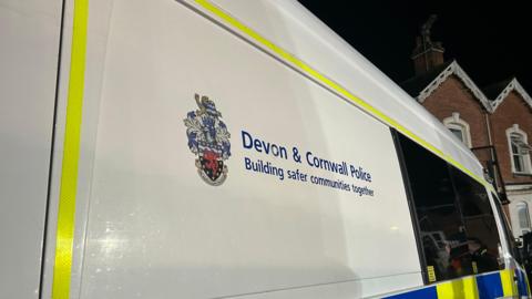 The side of a Devon and Cornwall Police vehicle. The van is white and has a yellow border.