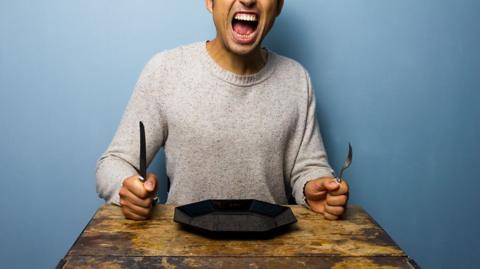 A person sitting at a table holding a knife and fork upright with their mouth wide open in anger.