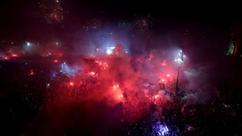 An night aerial shot showing crowds of people, some holding aloft lit flares.