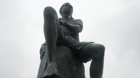 A low-angled shot of the Benjamin Britten statue. It depicts a 14-year-old boy sitting on what appears to be a rock. He is looking forwards. 