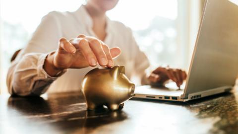 A woman's right hand touching a small golden piggy bank in the foreground. Her left hand is on a laptop keyboard in the background.