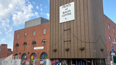 A large red brick building with multiple sections set back from one another and a ramp to the right side. There is a sign above the glass entrance doors which reads Prince of Wales Centre. The section to the left of the entrance has a number of archways containing colourful murals.