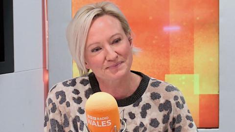 Ruth Dodsworth is sitting down and wearing a leopard print top. In front of her is a microphone, and behind her is a vertical digital screen, both of which are a bright orange. The microphone says BBC Radio Wales on it.