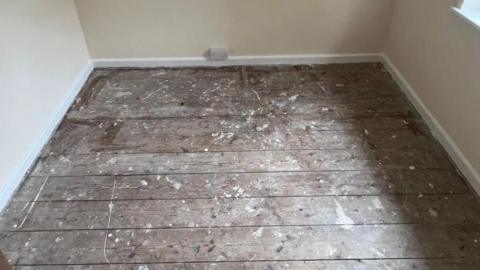 Empty room with bare floorboards. The wall is cream. There are white rims around the floorboard. There is an outlet on the wall. 