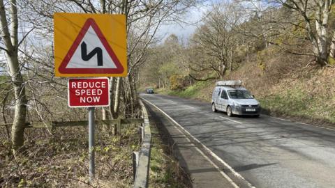 A country road with a van passing and a reduce speed sign