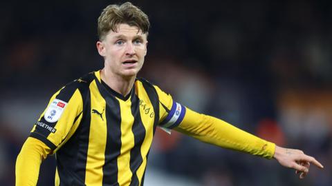 George Byers in action for Port Vale. He is wearing a yellow and black striped away shirt and is pointing out an instruction with his left arm.