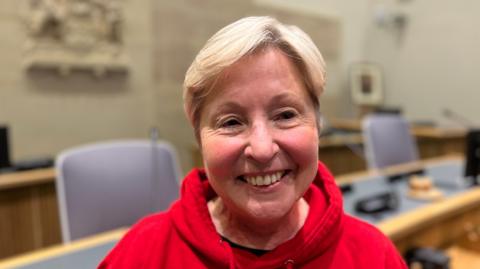 Valerie has short blonde hair and is smiling at the camera. She is wearing a red hoodie standing in a council focus. The benches and chairs behind her are out of focus.