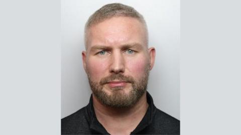 A mugshot of Konrad Labinski. He is wearing a black shirt. He has a neatly trimmed beard and moustache and short fair hair.