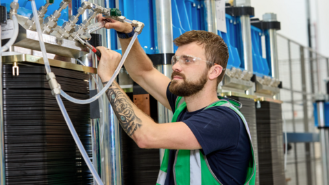 A man with a beard and wearing a green safety jacket and safety glasses uses a spanner to tighter a series of metal pipes