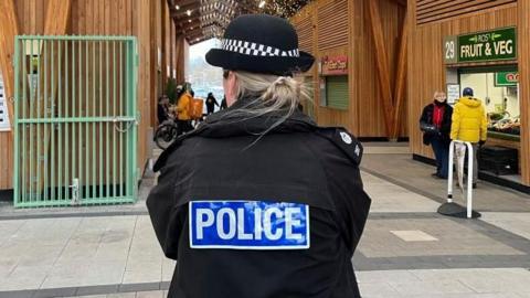 A female police officer who is wearing a police uniform and hat is standing inside Great Yarmouth's market building. She has her back turned to the camera.