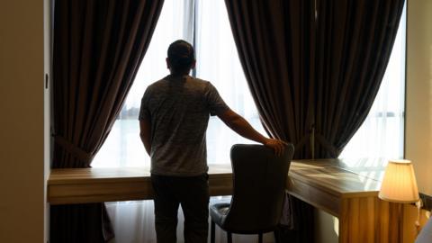 A man stares out of a hotel room window. The windows are large and surrounded by brown curtains. He puts his hand on a leather chair. There is an L-shaped desk and a lamp. 