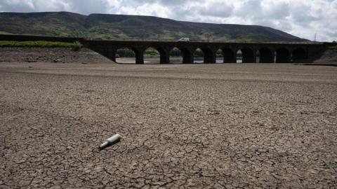 Parched, cracked land in the foreground. An empty bottle lies on the surface. In the background is a bridge with a white van driving across.