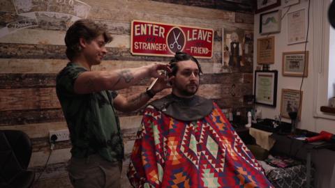 A barber with a green shirt and brown trousers stood and cutting the hair of a man who is wearing a patterned gown and sat in a chair having his hair cut in a barbershop.