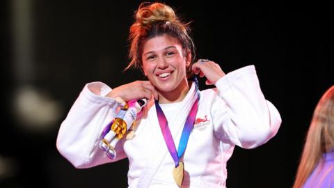 Acelya Toprak, wearing England's white judo robe, smiles with her gold medal around her neck on the podium