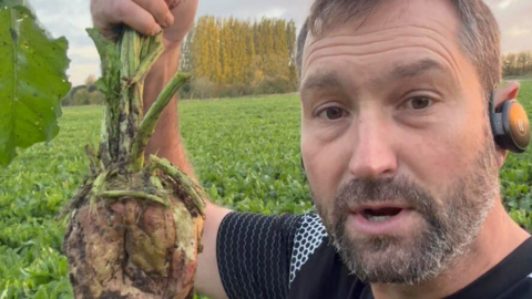 David Wheatley stands on a patch of open land, surrounded by earthy soil and growing sugar beets. He is dressed in a black top and has a black device on his ear - likely an earphone. The area is rural and practical, with no buildings in sight, just the land sugar beets are growing. He is holding one up to his face by the long green leafy stem. The sugar beet itself is a diamond shape with soil on it. Mr Wheatley  has brown hair and facial hair that is greying slightly. 
