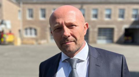 A head and shoulder shot of a man standing outside with a building and the back of a red and yellow fire engine behind him. He has a bald head and facial hair and is looking into the camera. He's wearing a grey suit over a white shirt, and is wearing a grey tie.