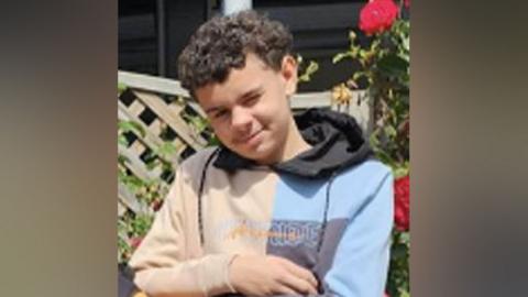 Darrian Williams. He had short curly dark hair and is wearing a blue and beige hoodie. Behind him is a garden trellis and pink roses. He is smiling at the camera.