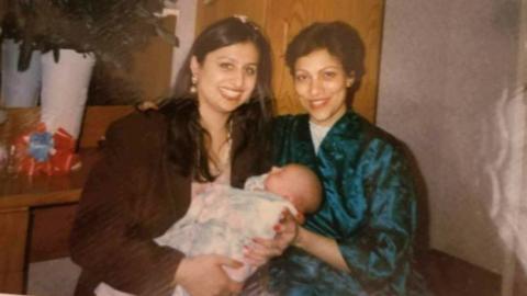 The image shows two women holding a baby. The woman on the left is young with long black hair while the woman on the right is older with short black hair.