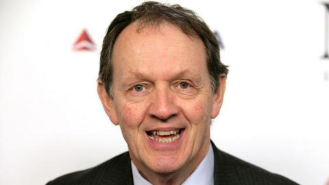 Kevin Whately standing in front of a white background. He is wearing a suit and is clean shaven. He has dark hair and is smiling at the camera. 