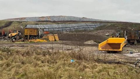 A view of the landfill site in Pilsworth on the border of Bury and Rochdale. The land is quite barren and three dumper trucks or trailers towed by tractors are in the background.