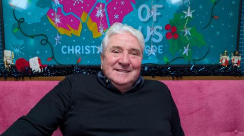 John Linehan sitting on a pink sofa smiling at the camera with some Christmas decorations and the branding of The State of Us podcast in the background. He is wearing a navy jumper and a blue shirt.