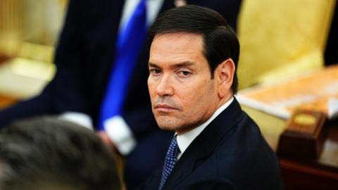 Marco Rubio, with a black suit and blue tie, looks left towards the camera 