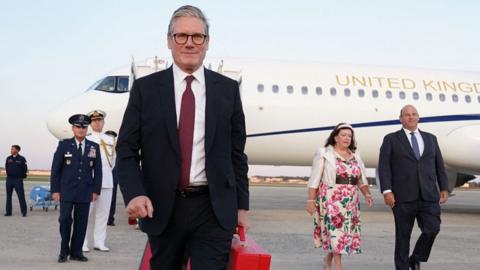 Starmer walks in a suit and tie and carrying a red briefcase, with his private jet in the background that says 'United Kingdom', and members of crew and his team also in the background, after landing in Maryland on a trip to Washington DC on 12 September