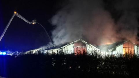 The warehouse at the garden centre. An aerial ladder is being used to tackle the blaze. Smoke and fire can be seen.