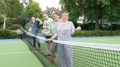 Four people are pictured holding blue tennis rackets and a tennis ball each while standing by a tennis net.