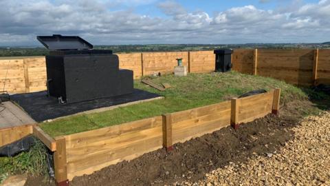 The black-painted concrete entrance to the bunker stands on a grass mound, with wooden edging on the side and a fence around the perimeter.