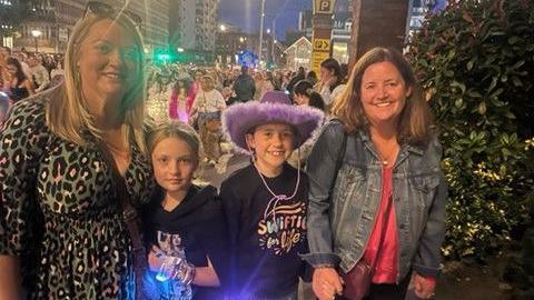 two mum standing either side of their young daughters wearing concert bracelets, all standing in a crowd on the street 