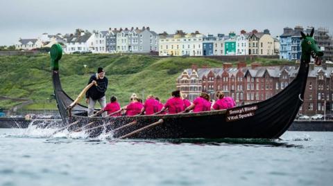 A team of women in pink T-shirts row a boat while the coxswain stands at the stern of the boat holding the rudder. Peel can be seen in the background.